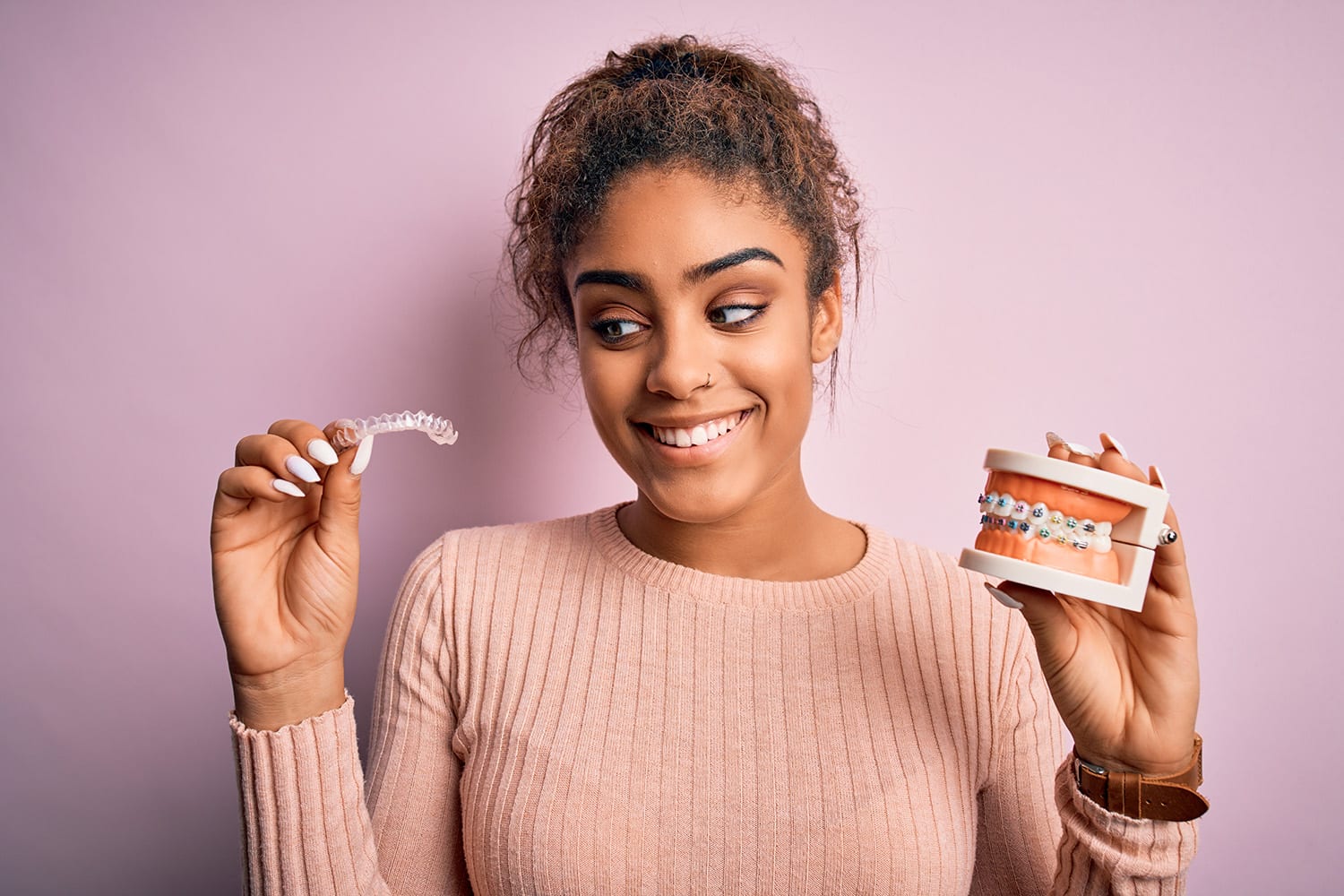Smiling patient holding a clear Invisalign aligner in one hand and a traditional metal braces model in the other